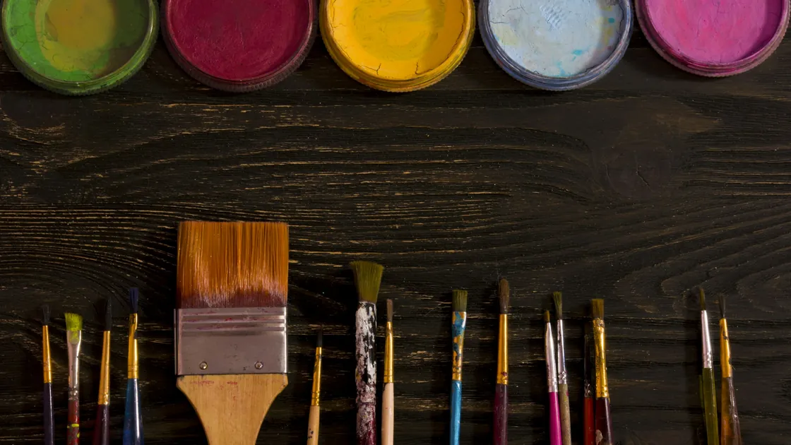 Pinceles de distintas formas y tamaños alineados sobre un tablero de madera oscura junto a botes de pintura abiertos